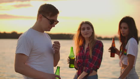 Female-students-are-dancing-with-beer-on-the-hot-summer-party-on-the-beach-with-their-boyfriend-in-white-T-shirts.-They-enjoy-this-summer-evening-at-sunset.
